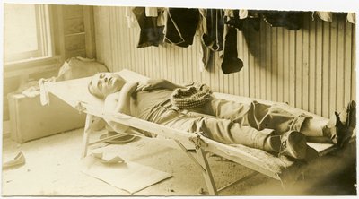 Fotografia orizzontale seppia che mostra un uomo che dorme su una branda in un edificio con pannelli di legno. Vari articoli di abbigliamento possono essere visti appesi al muro e valigie possono essere viste a terra nell